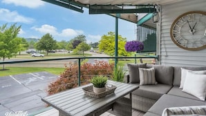 A covered outdoor seating area with a wicker sofa, a wooden table with a plant, and a large clock on the wall.