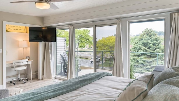 A bedroom with a large bed, a desk, a chair, a television, and a view of a balcony with a railing and greenery.