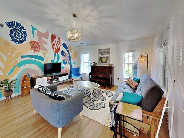 Una sala de estar con una televisión de pantalla plana, un sillón azul, un sofá gris, una mesa de centro de madera y un mural en la pared.