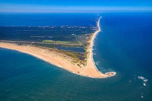An aerial view of a coastal area with a long stretch of beach, a small peninsula, and a large body of water.