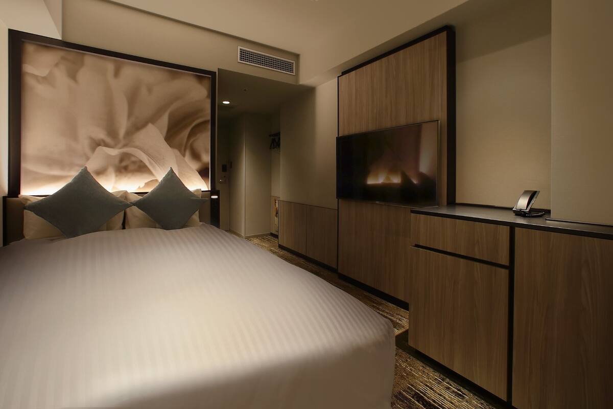 A modern hotel room with a large bed, a flat-screen TV mounted on the wall, and a wooden wardrobe.