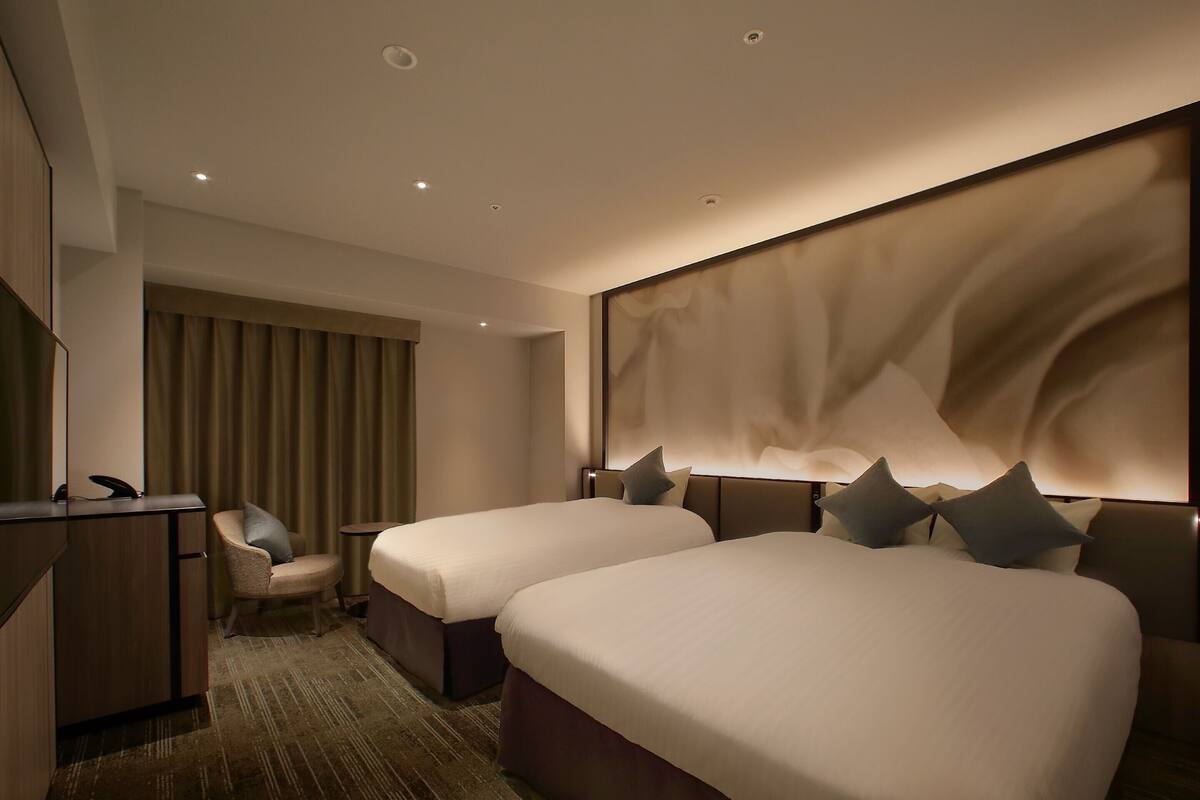 A hotel room with two beds, a desk, a chair, and a large wall-mounted light fixture.