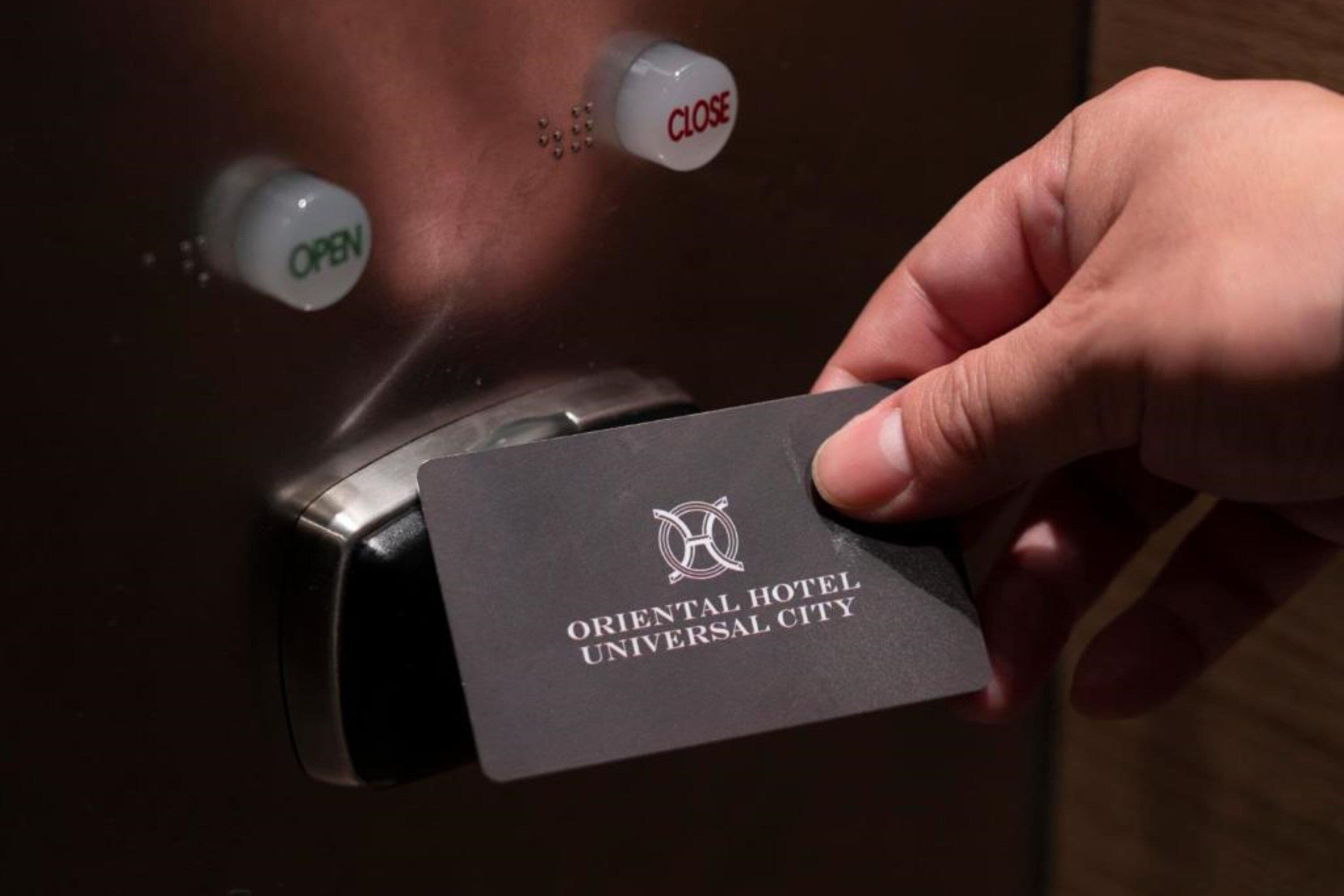 A hand holding a card with the text "ORIENTAL HOTEL UNIVERSAL CITY" in front of a door with an "OPEN" sign and a "CLOSE" sign.