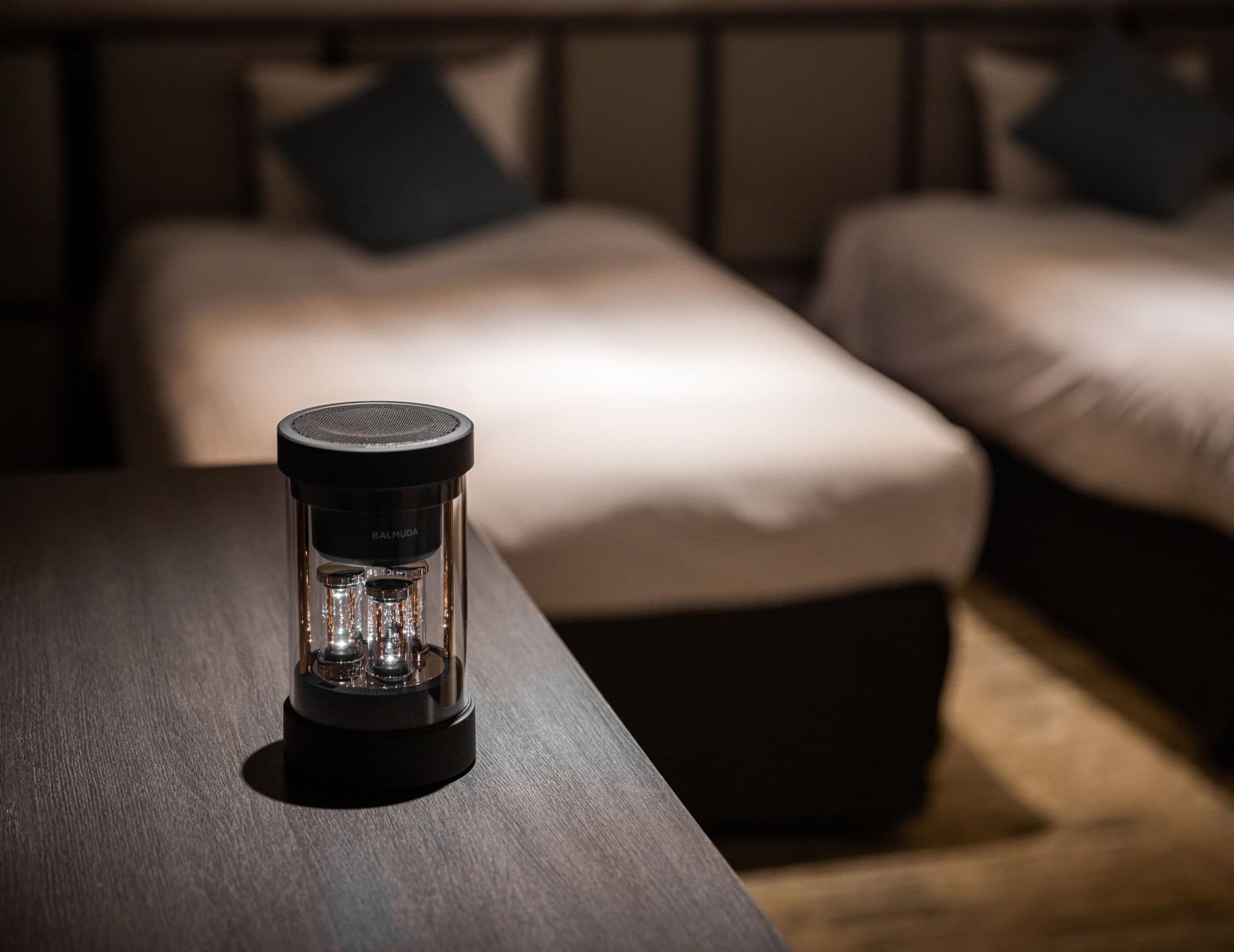 Gros plan d’un objet cylindrique posé sur une surface en bois, avec un arrière-plan flou représentant un lit et des oreillers.
