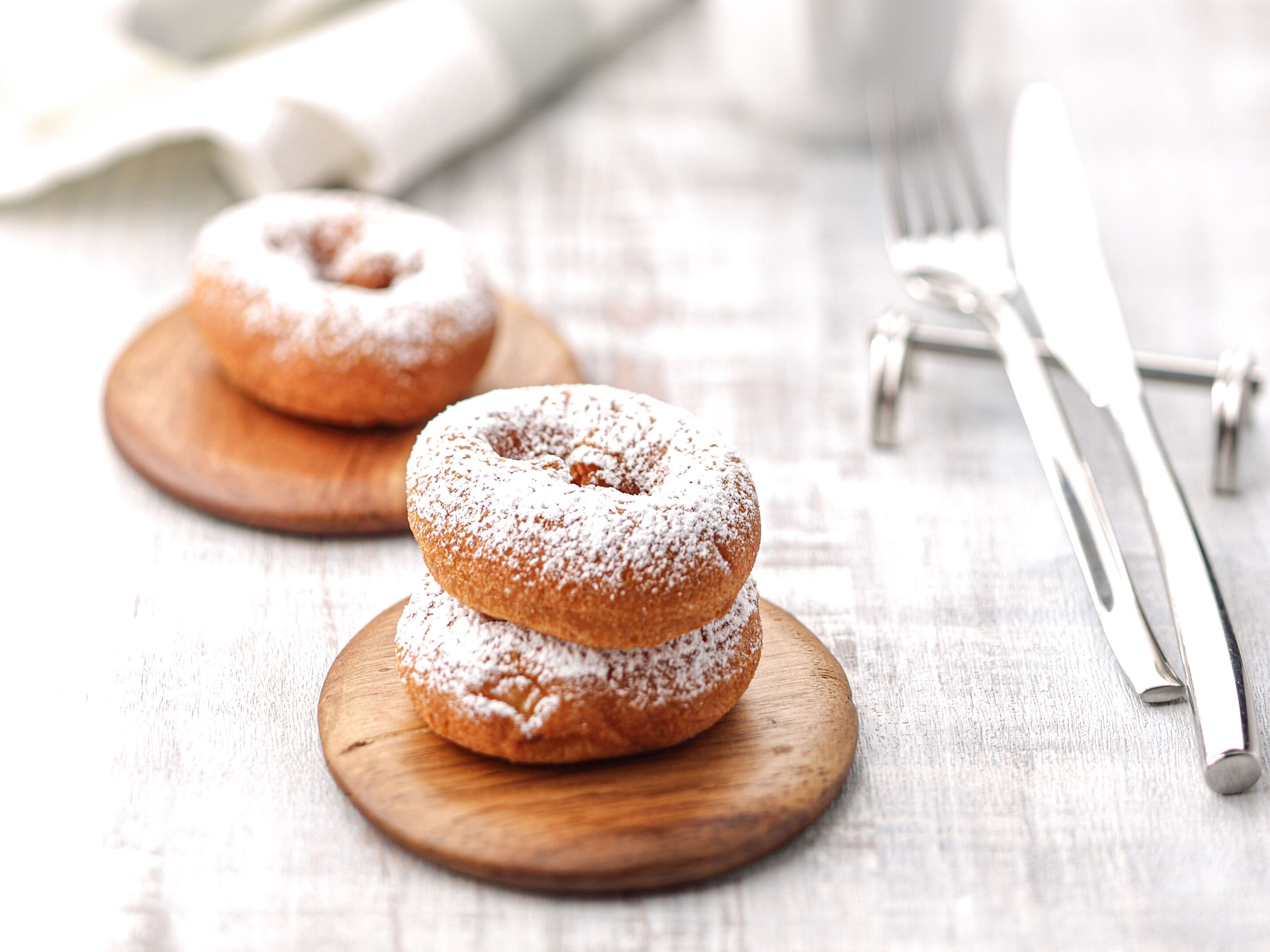 Two powdered donuts on wooden plates with a fork and knife beside them.