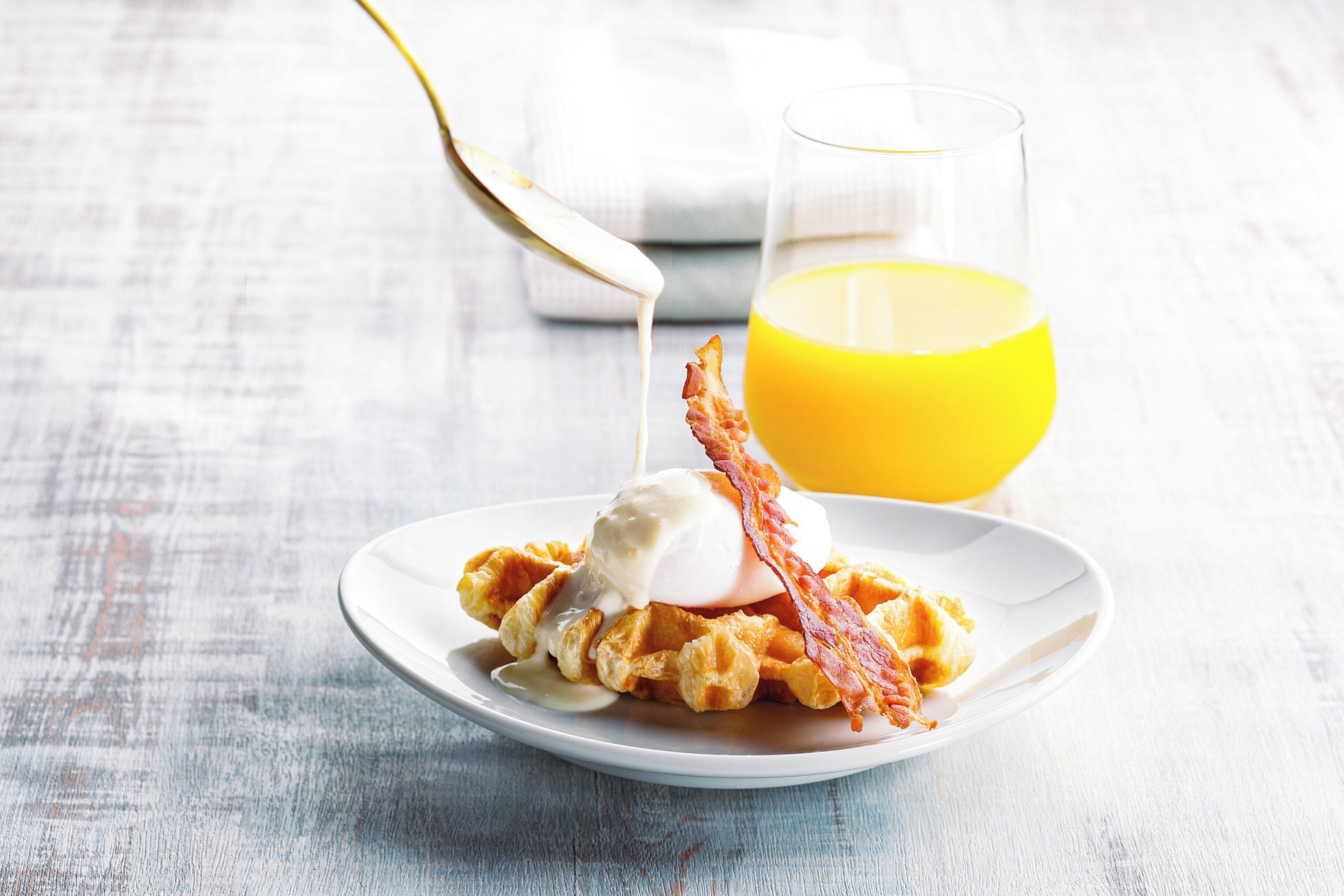 Un plato de waffles cubiertos con un huevo pochado y tocino, acompañado de un vaso de jugo de naranja.