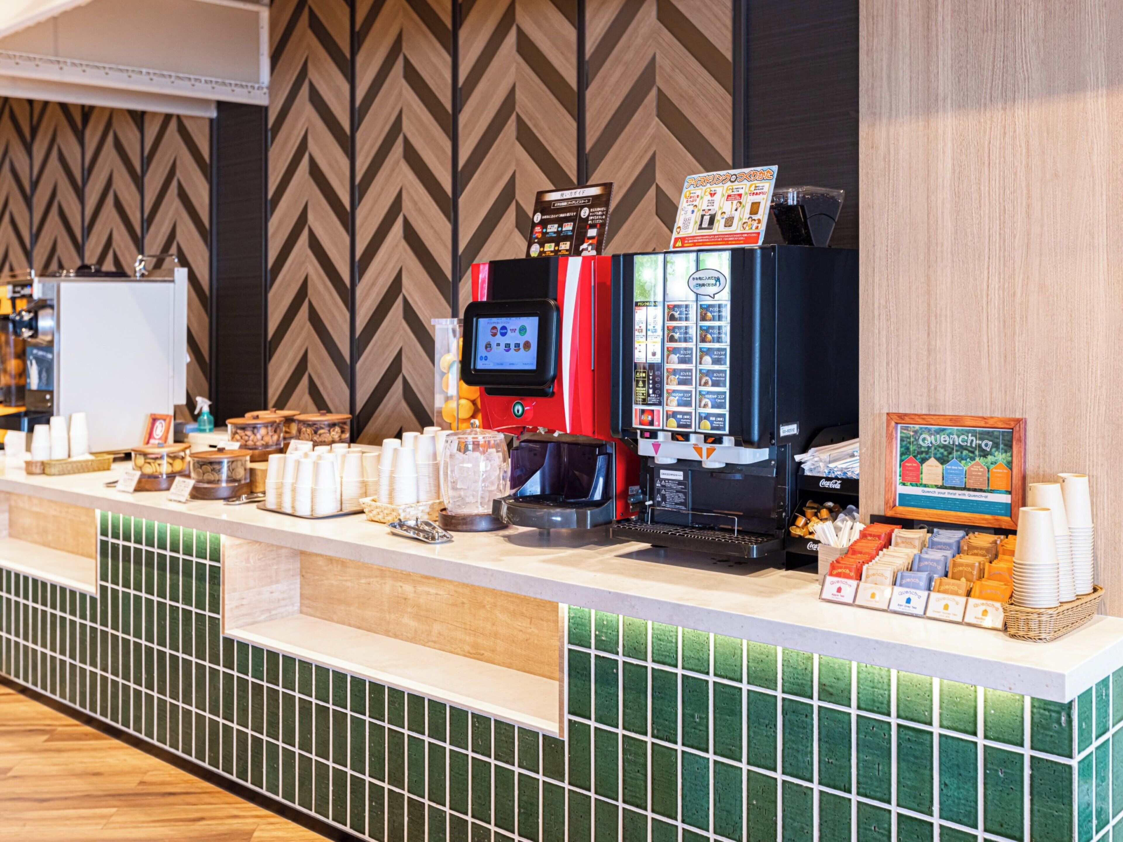 Estación de café con varias máquinas de café, dispensador de Coca-Cola y una variedad de cápsulas de café.