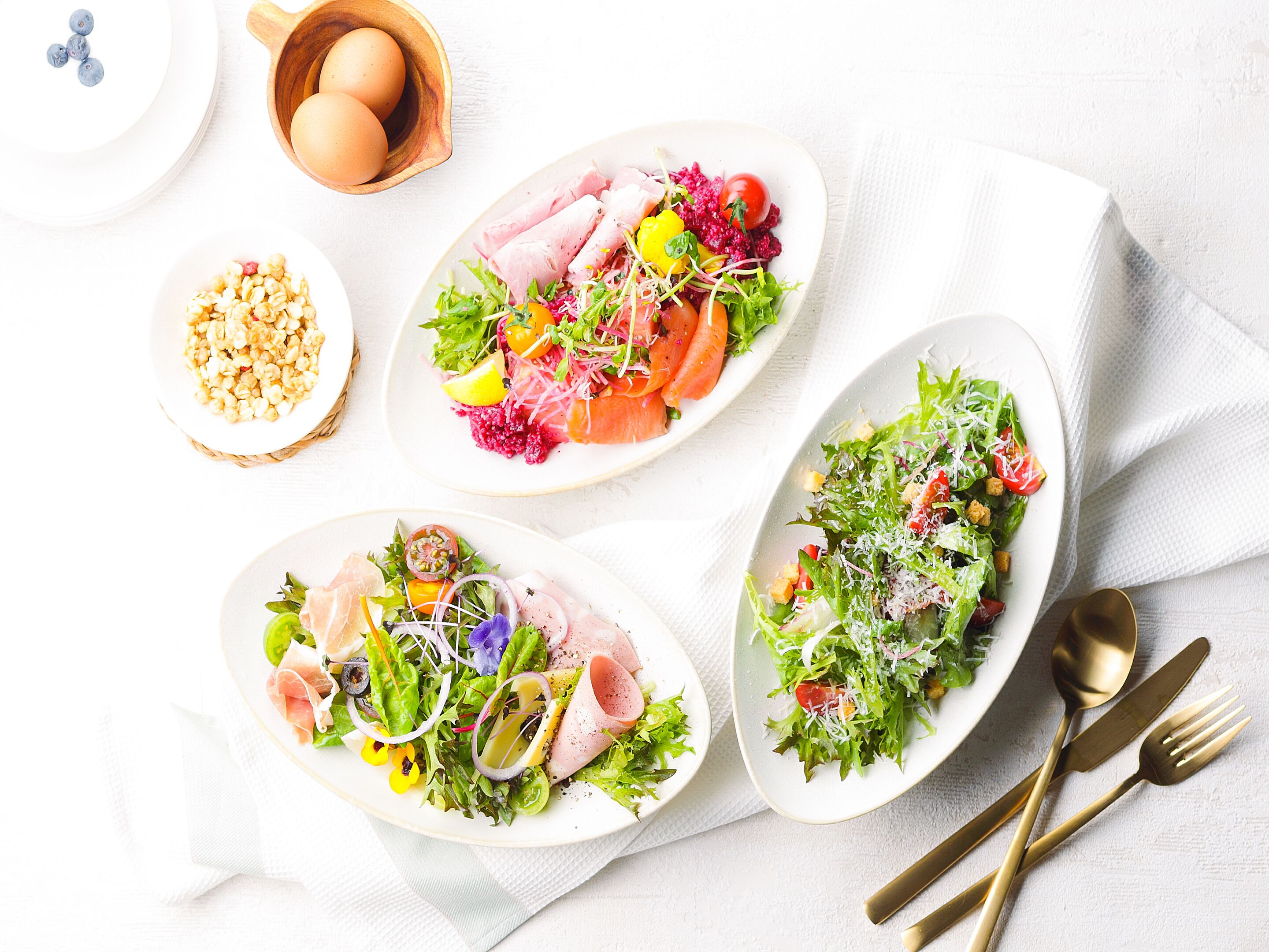 Tres platos de comida con diversos ingredientes y guarniciones, acompañados por un bol de frutos secos, un cuenco de madera con huevos y una servilleta con cubiertos sobre un mantel blanco.