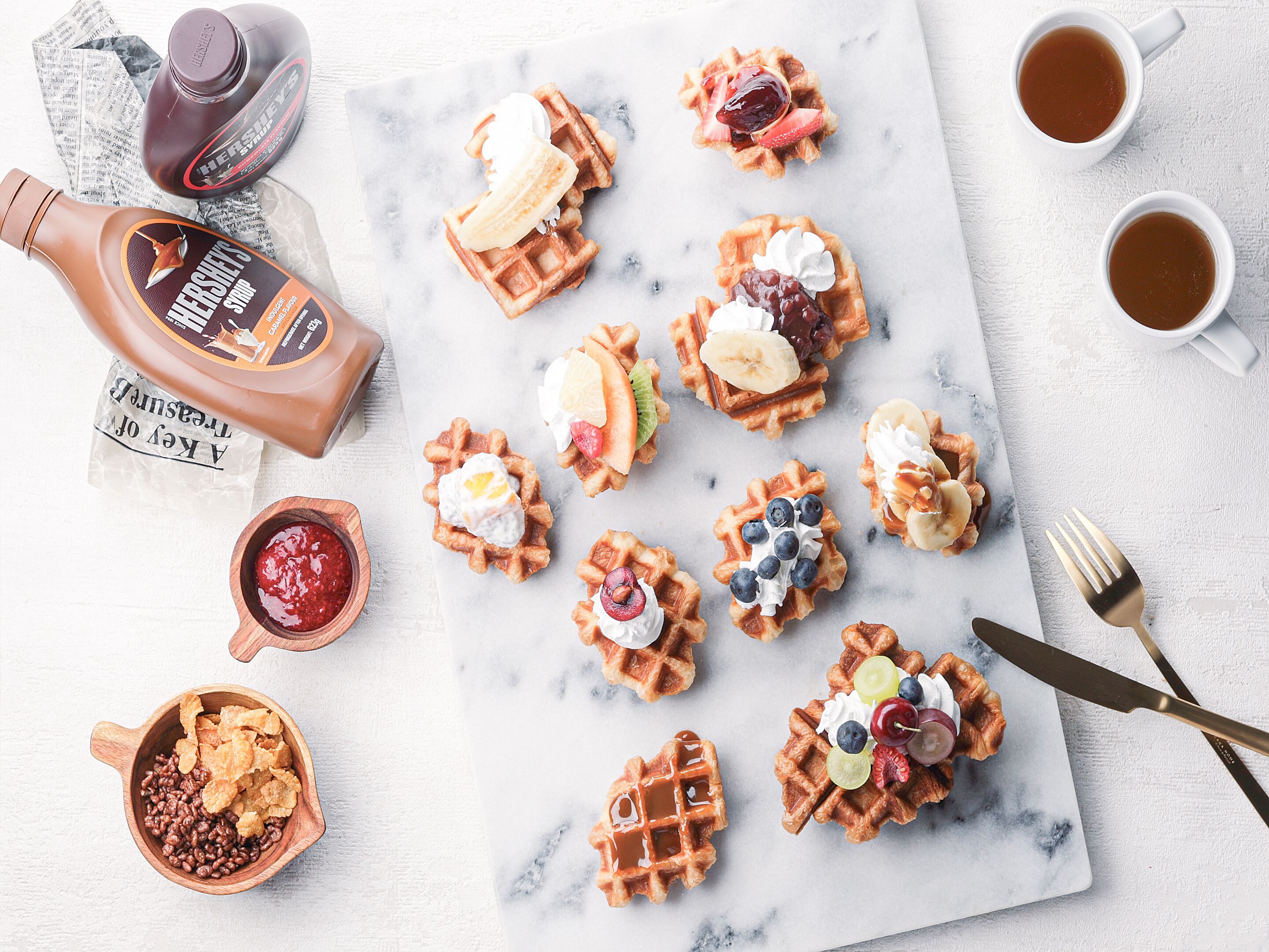 A variety of waffles with different toppings, a bottle of Hershey's chocolate syrup, a bowl of chocolate chips, and two cups of coffee on a marble surface.