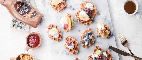 A variety of waffles with different toppings, a bottle of Hershey's chocolate syrup, a bowl of chocolate chips, and two cups of coffee on a marble surface.
