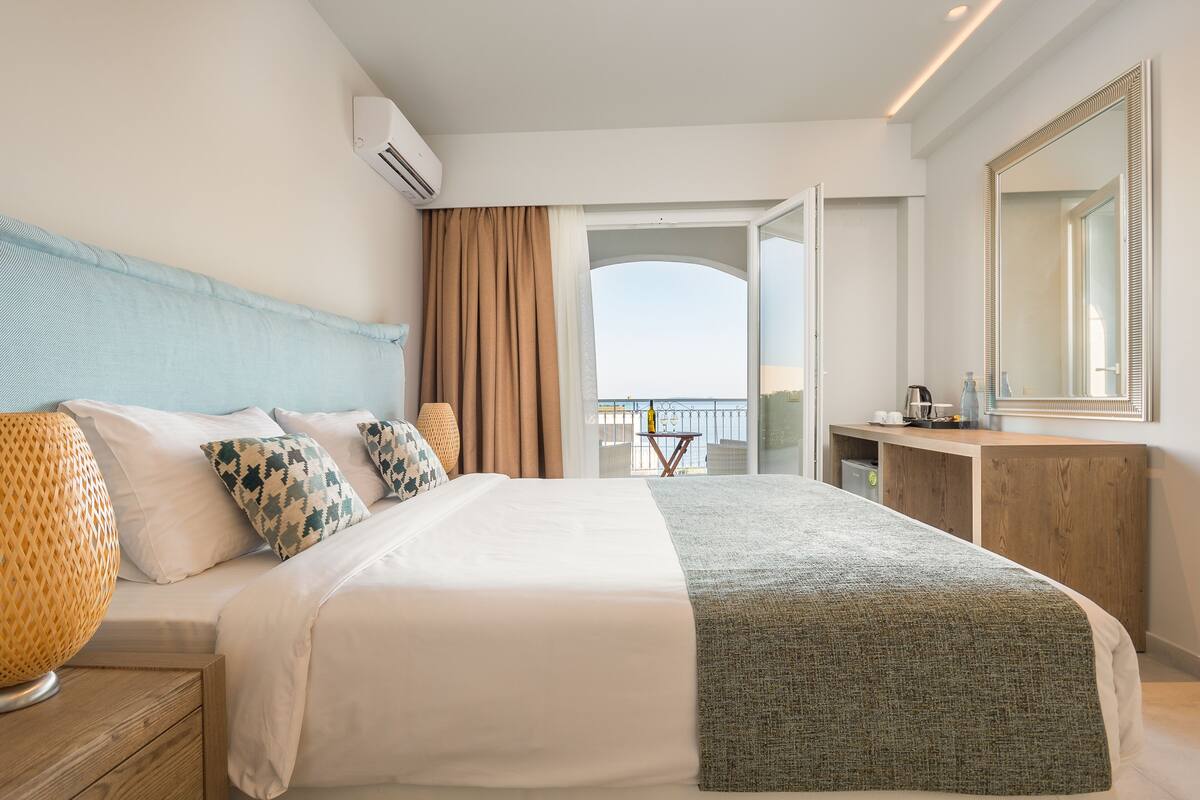 Quarto de hotel com uma cama grande, uma escrivaninha, um espelho e vista para o mar.