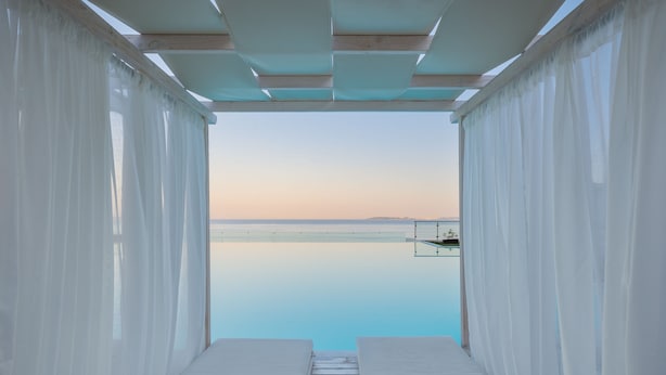 Vista desde una habitación con dos camas blancas, suelo de madera y cortinas blancas, con vistas a una masa de agua tranquila y una costa distante.
