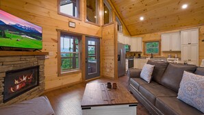 A cozy cabin interior with a stone fireplace, a large flat-screen TV, a brown leather sofa, and a kitchen area.