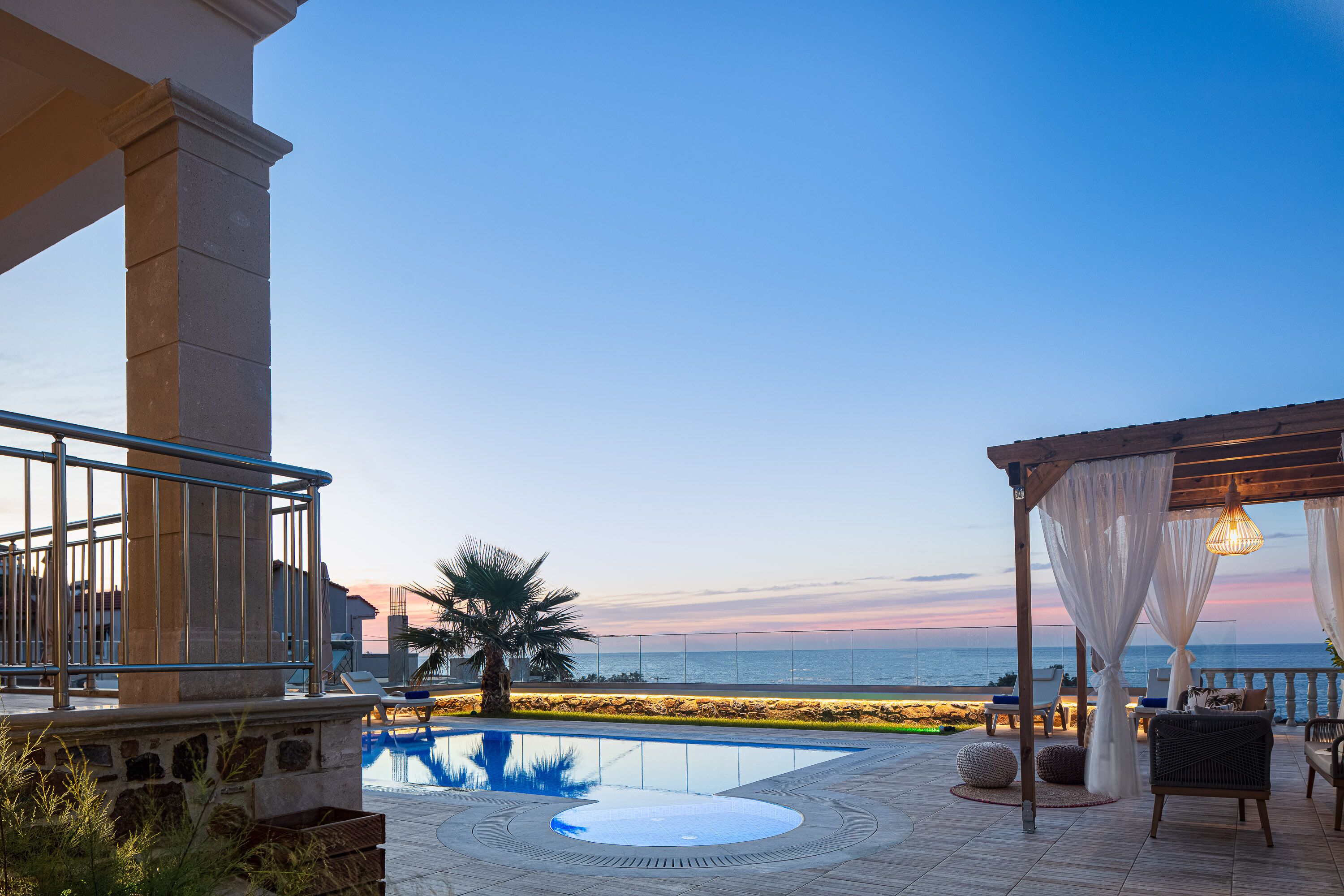 A pool area with a view of the sea, a pergola with white curtains, and lounge chairs.
