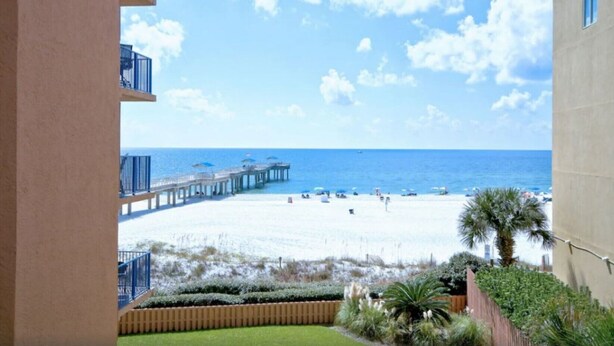 A beach view from a balcony with a pier extending into the ocean, a sandy beach, and some greenery.