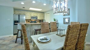 A dining area with a light-colored table and wicker chairs, a hanging light fixture, and a kitchen in the background.