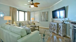 A living room with a sofa, coffee table, and a television. There is a ceiling fan and a sign that reads "BEACH HOUSE".