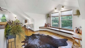 Ein geräumiges Zimmer mit großem Fenster, Holzboden, Kuhfellteppich und einer Sitzecke mit Bank und Kissen.
