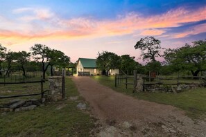 A rural property with a dirt path leading to a house with a green roof, surrounded by trees and stone walls.