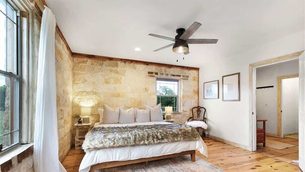 A bedroom with a stone wall, wooden floor, a bed with a fur throw, a ceiling fan, and a window with curtains.
