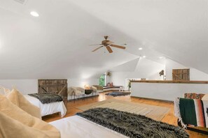 Um quarto amplo no estilo loft, com teto inclinado, ventilador de teto e uma grande janela que permite entrada de luz natural.