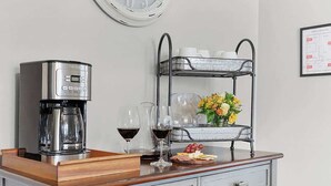 Una cafetera, copas de vino, una bandeja de frutas y un estante metálico con tazas y flores sobre una encimera de madera.