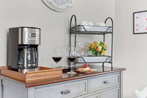 Una cafetera, copas de vino, una bandeja de frutas y un estante metálico con tazas y flores sobre una encimera de madera.