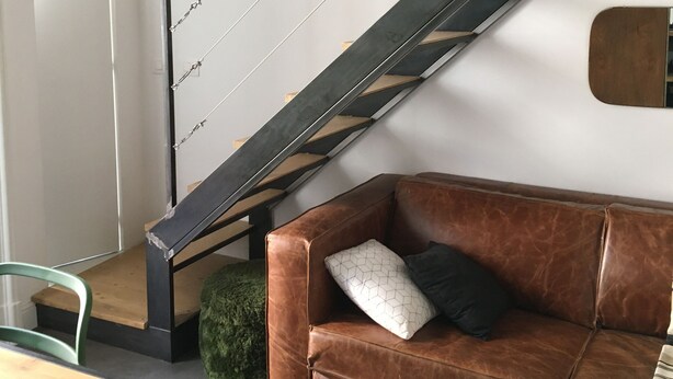 Un salon moderne avec un canapé en cuir marron, une table basse en bois et un escalier doté d’une rampe en métal.
