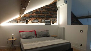 Une chambre avec un mur en pierre, un lit, une petite table avec une lampe et un radiateur.