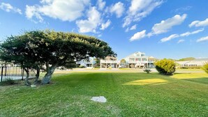 A well-maintained lawn with a large tree and a building with multiple windows and a porch in the background. - DOG FRIENDLY - Second Row ...Ruff Life beach cottage. (Oak Island)