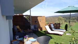 Een terras buiten met een eettafel, twee lounge stoelen en een houten terras.