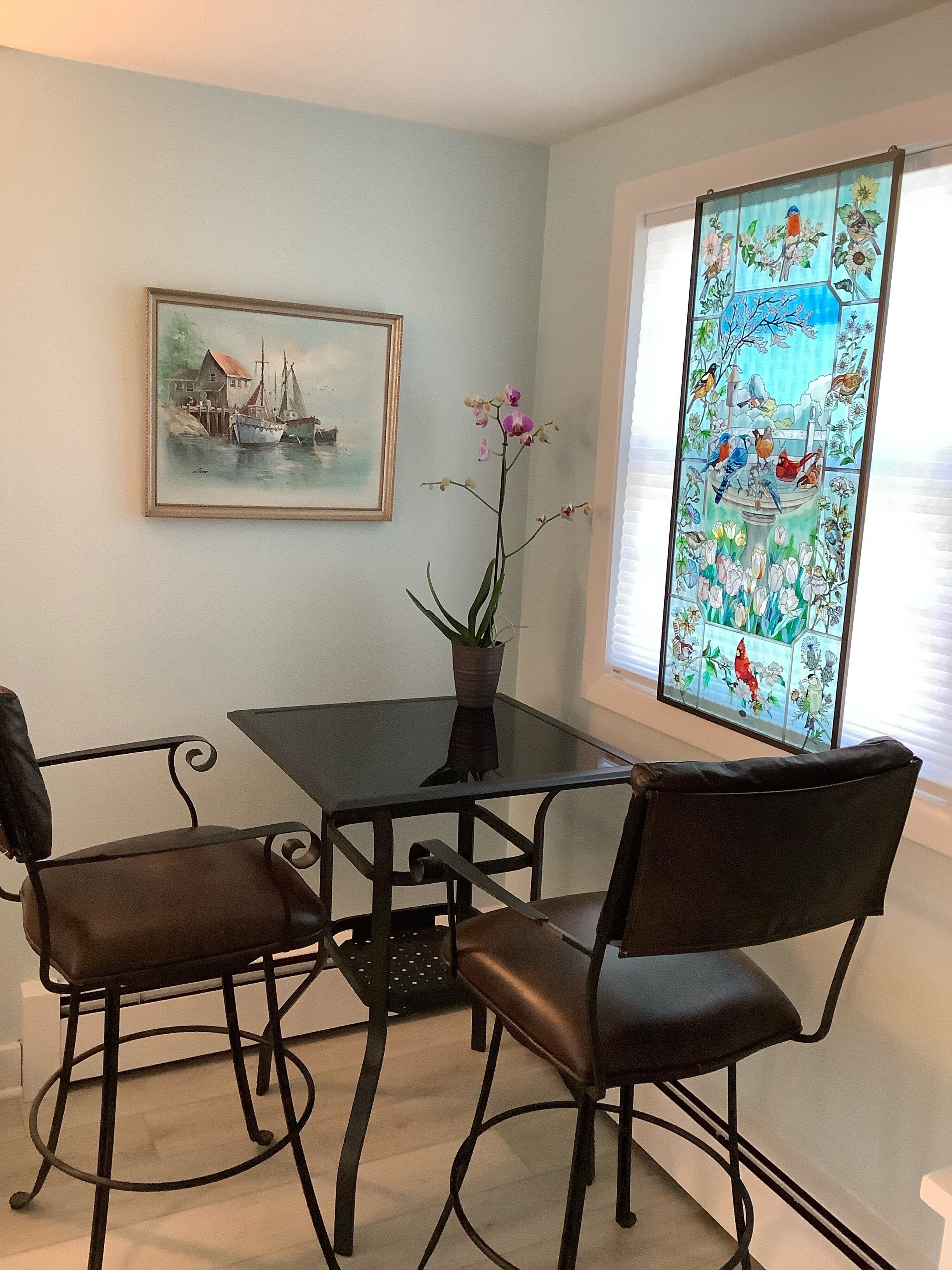 A dining area with a glass table and four chairs, a potted orchid, a framed painting, and a stained glass window.