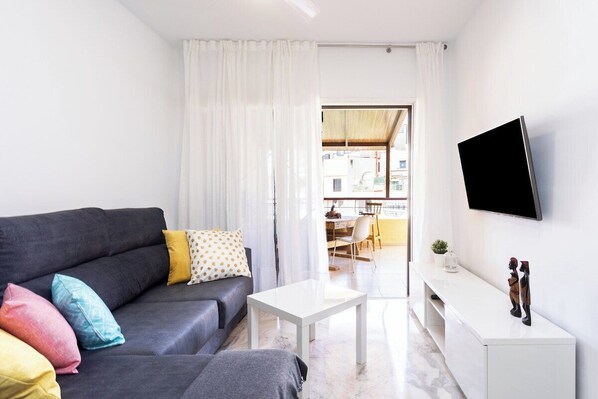 Ein modernes Wohnzimmer mit einem grauen Sofa, bunten Kissen, einem weißen Couchtisch und einem flachbildfernseher an der Wand.