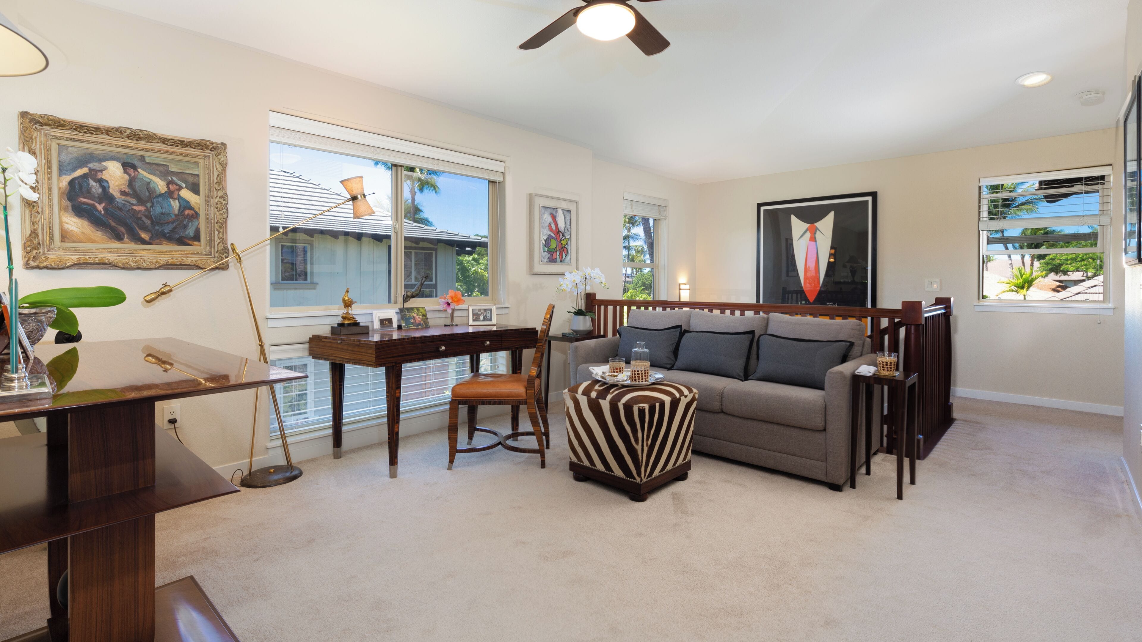 A living room with a sofa, a desk, and a chair. There are windows with blinds, a ceiling fan, and a framed picture on the wall.