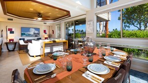 A dining area with a large table set for six, a kitchen in the background, and a view of the outdoors. - Sophisticated freshly remodeled 3bd+/3ba, two story villa w/ Private Beach club (Puako)