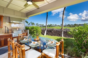 An outdoor dining area with a glass table, wooden chairs, and a view of a lush landscape with palm trees and a clear sky.