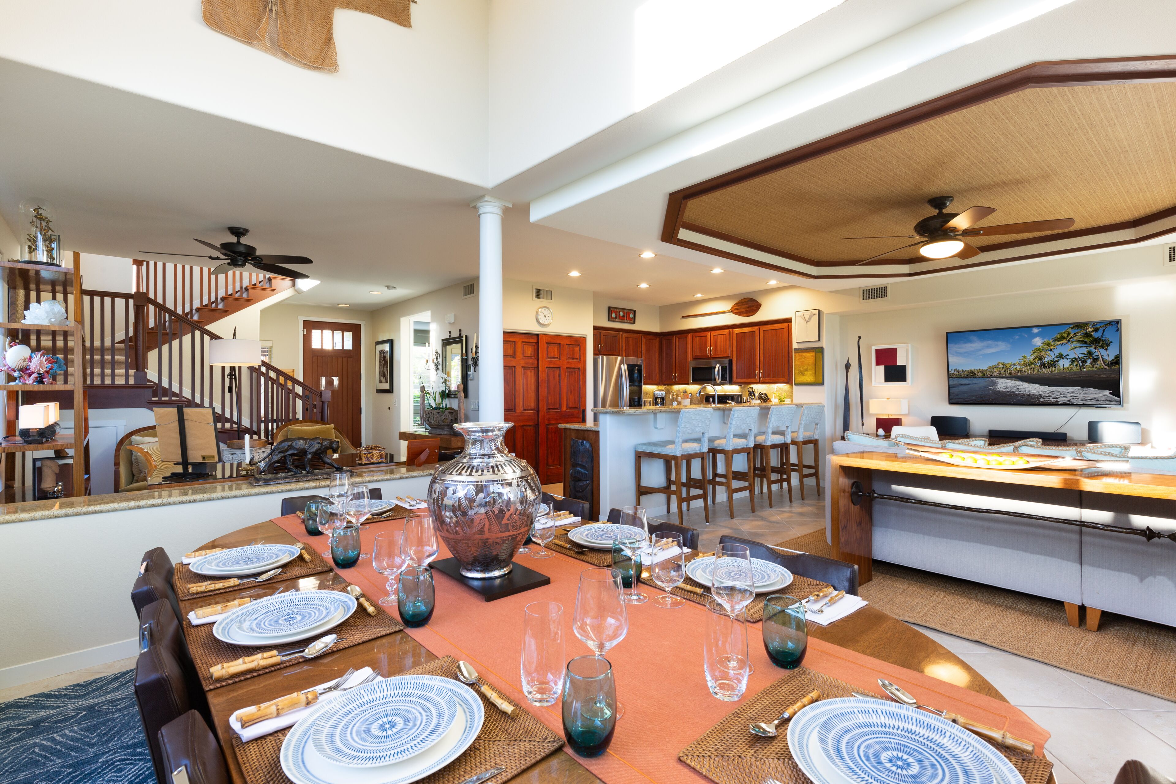 Un comedor espacioso con una gran mesa dispuesta para una comida, una cocina al fondo y una sala de estar con televisor.