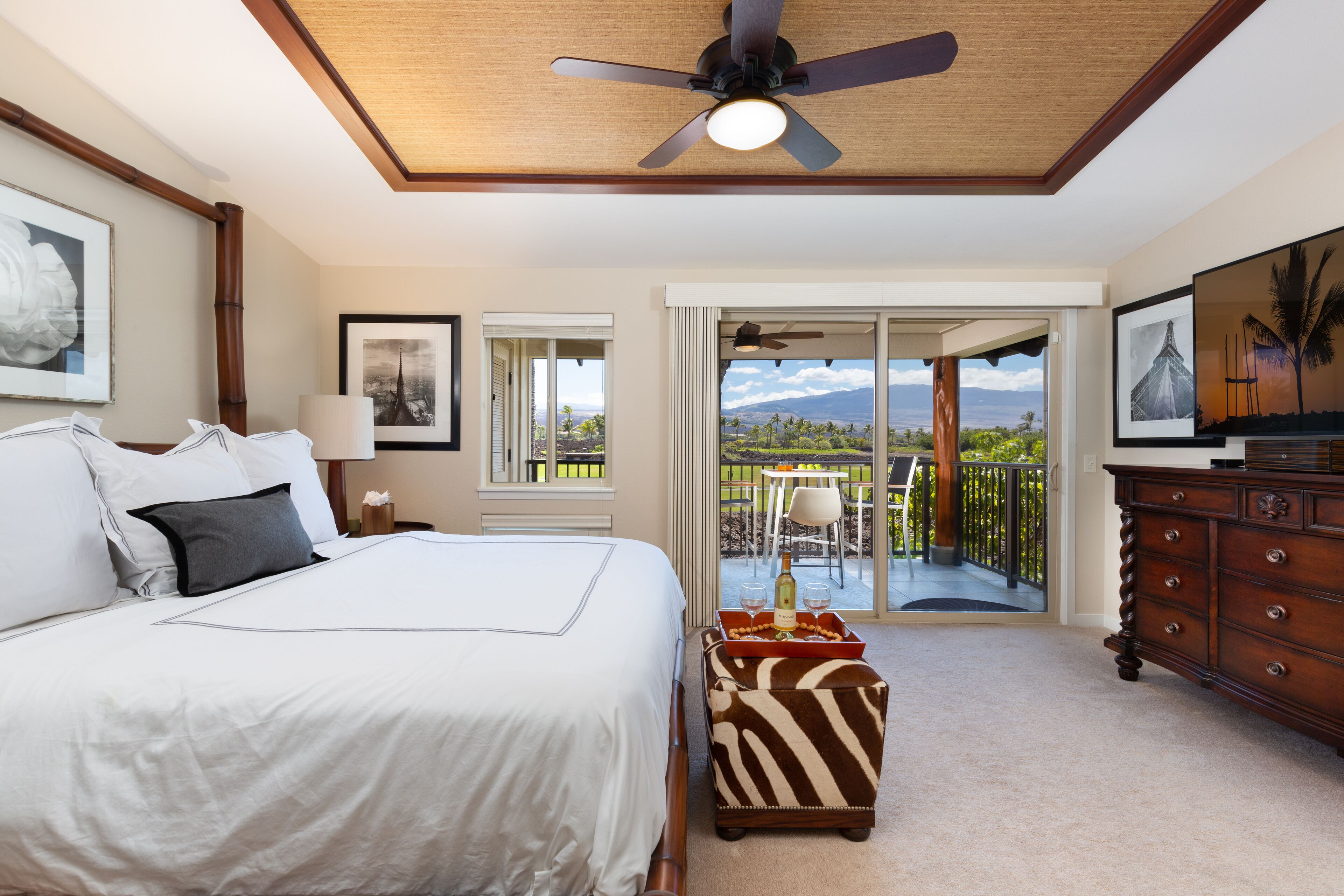 A bedroom with a bed, a dresser, a ceiling fan, and a balcony with a view.