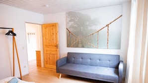 A room with a grey sofa, wooden floor, a wooden door, and a rope bridge painting on the wall. - Ferienwohnung mit Seeblick Sport und Erholung (Winterberg)