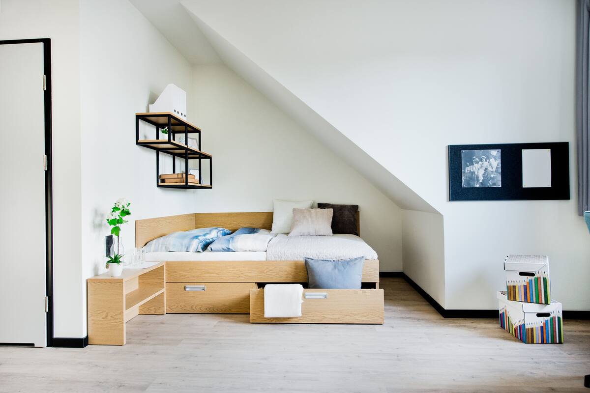 Een moderne slaapkamer met een schuine plafond, een houten bed met opbergladen, een nachtkastje met een plant en een wandplank.