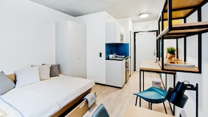 A modern hotel room with a bed, a desk, a chair, a small kitchenette, and a bookshelf. - YOUNIQ Gdansk - Campus Accommodation (Gdansk)