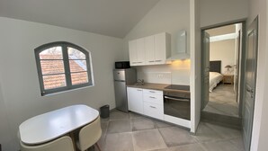 A compact kitchen with white cabinets, a microwave, and a stove. There is a small dining area with a table and chairs. A window provides natural light, and a door leads to a bedroom.