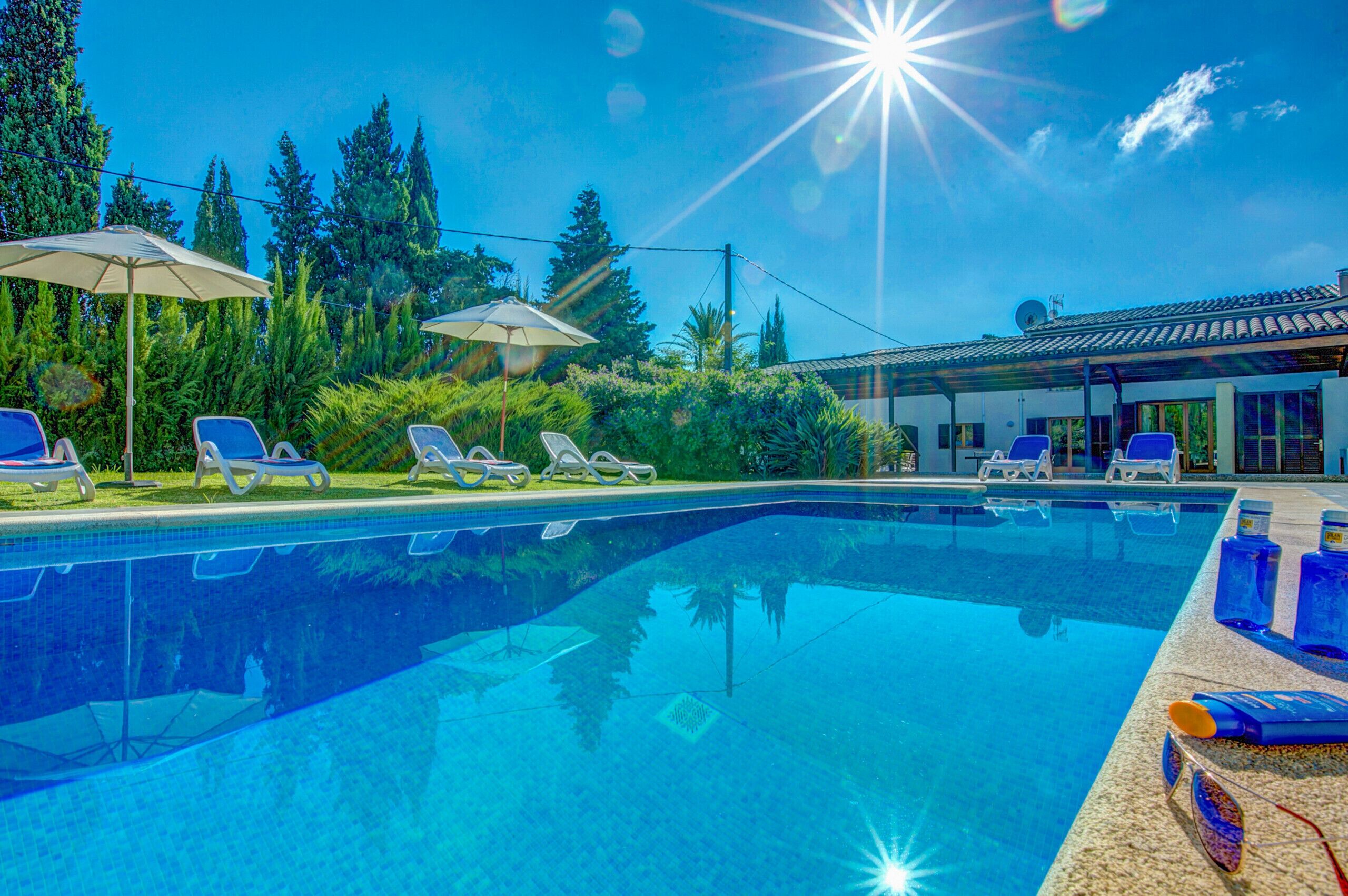 Piscina azul límpida com espreguiçadeiras e guarda-sóis, rodeada por vegetação e um edifício com telhado em azulejos.