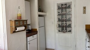 Cocina compacta con armarios blancos, un refrigerador pequeño, una estantería de madera con una botella y una puerta de vidrio decorativa.