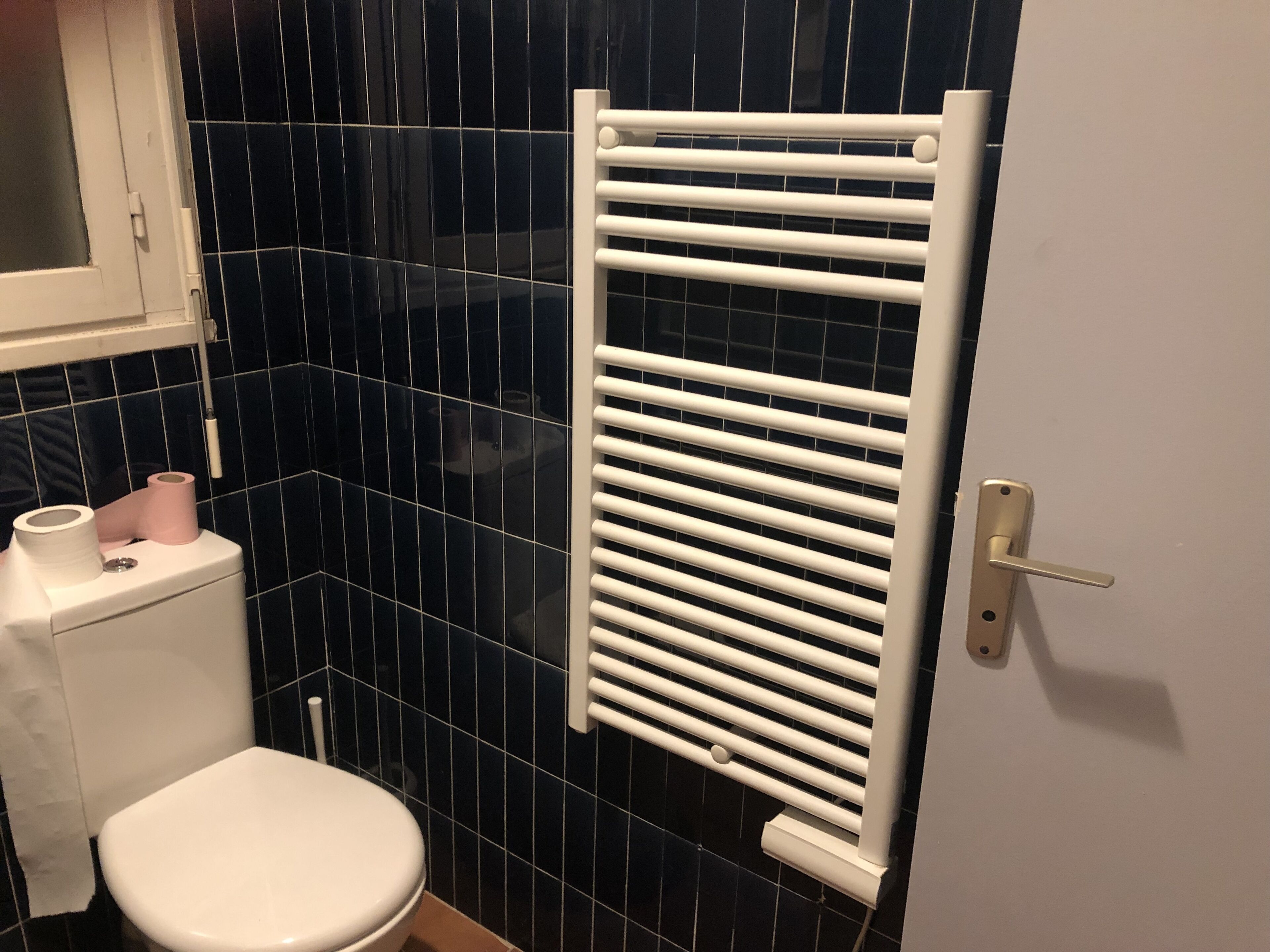 Un baño con inodoro blanco, paredes revestidas de baldosas negras, radiador blanco y una puerta con manija.