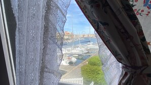 Uitzicht door een raam met kantgordijnen, waarop een jachthaven met boten en een reuzenrad op de achtergrond zichtbaar is.