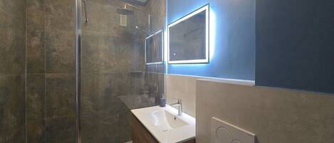 Un baño moderno con cabina de ducha de vidrio, un lavabo blanco y un espejo con iluminación perimetral.