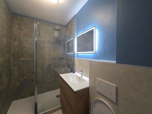Un baño moderno con cabina de ducha de vidrio, un lavabo blanco y un espejo con iluminación perimetral.