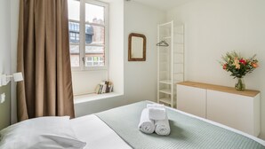 A hotel room with a bed, a window with curtains, a white wardrobe, a mirror, a shelf with books, and a vase with flowers. - Le Moulin des Carmélites 1 - Appt moderne à Rennes (Rennes)