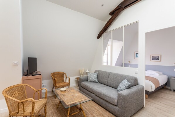 Une chambre avec un canapé gris, deux fauteuils en osier, une table basse en verre, un lit avec une literie blanche et un grand miroir.